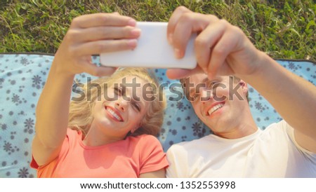CLOSE UP: Cheerful couple laughing and taking selfies while lying on a comfortable blue blanket. Happy man and woman having fun taking photos of themselves during an outdoor date in tranquil nature.
