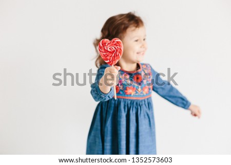 Little girl eating red heart shape lollipops wearing blue jeans dress on light background