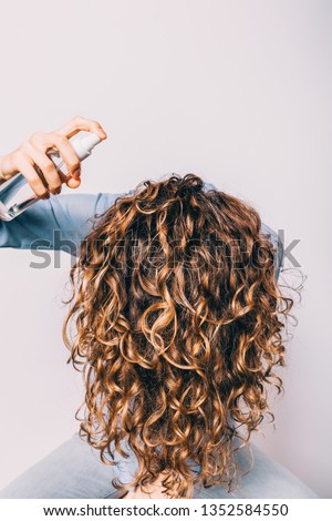 Head of young woman with long curly hair is lowered down to apply cosmetic care product. Female using spray with sea salt to make beachy waves hairstyle.