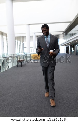 Front view of happy young African-American businessman with coffee cup using mobile phone while walking in modern office. He is smilling