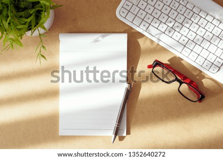 Top view of office arrangement on wooden desk. Empty paper page from notepad with pen, eye glasses, white computer keyboard and green plant.