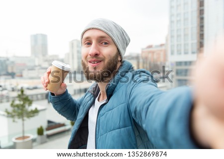 Happy young man giving a selfie portrait to his smart phone. He is holding a paper cup with hot coffee or tea in his hand while standing against the background of the city.