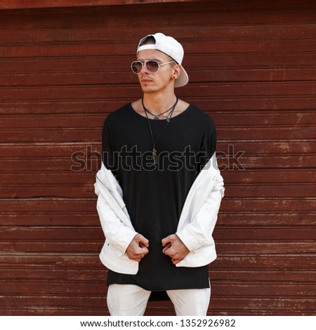Handsome hipster young man in a fashionable white summer jacket in a stylish black t-shirt in a trendy baseball cap in dark sunglasses near a wooden vintage wall. Urban stylish guy model.Men's fashion