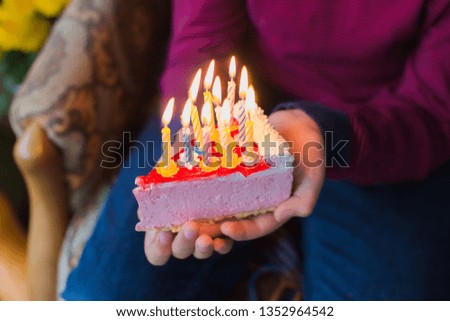 Closeup top view of hands of kid holding piece of birthday cake with burning colorful candles. Horizontal color photography.