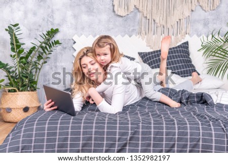Little beautiful daughter lies on her mother back, which looks at the tablet while lying on the bed