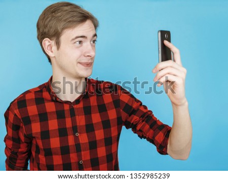 young emotional male teenager in red shirt on blue background looks into smartphone