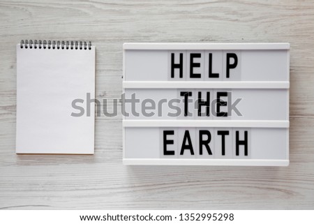 Modern board with 'Help the Earth' words, blank notepad over white wooden background, top view. Copy space.