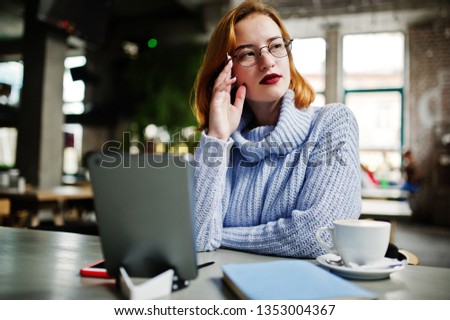 Cheerful young beautiful redhaired woman in glasses using her phone, touchpad and notebook while sitting at her working place on cafe with cup of coffee.