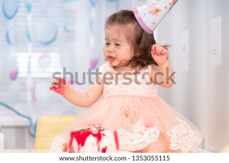 Little girl enjoying her birthday party sitting in front of the cake with sticky hands and face wearing a hat and pink dress