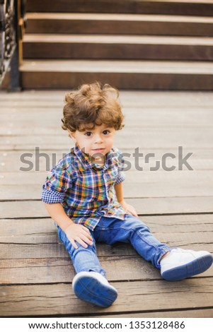 Portrait of handsome little toddler boy playing at summer city park. Calm child with beautiful curly brown hair and big eyes posing for photo sitting on wooden steps. Vertical color photography.