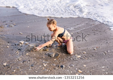 Having fun and joy concept. Blonde lovely girl with two braids playing with sea sand. Playful active kid on beach with waves in summer vacation.