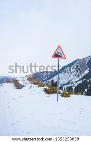 Road sign of dangerous descent in the mountains. Sign a steep descent in the winter mountains. Twelve percent on a road sign warning of a steep descent. Snowy mountain road.