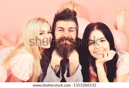 Man with beard and mustache attracts blonde and brunette girls. Girls fall in love with bearded macho, pink background. Threesome on smiling faces lay near balloons. Alpha male concept.