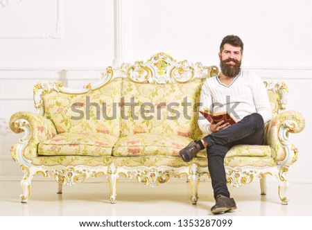Man with beard and mustache sits on baroque style sofa, holds book, white wall background. Guy reading old book with enjoyment. Reader concept. Macho on smiling face passionate about reading book.