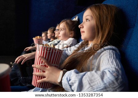 Pretty, little girl holding popcorn bucket, sitting with friends in cinema, in comfortable chairs. Children watching cartoon or movie. Leisure, entertainment.