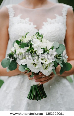 Young beautiful girl in elegant dress is standing and holding hand bouquet of white flowers and greens with ribbon at nature. The bride holds a wedding bouquet outdoors.