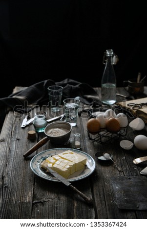 Rustic dark table with vintage plate of butter, strainer with flour, salt, sugar, whisk, spoons,  eggs, old cooking book, glasses of water. Process of cooking french dessert eclairs. Dark food photo.