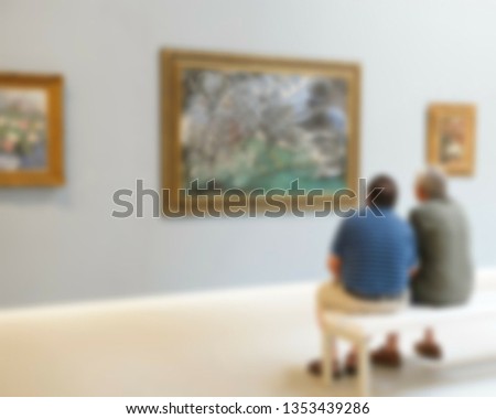 Two visitors sit on a bench and look at paintings in one of halls of Art Museum. Blurred view
