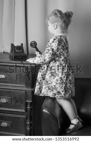 A beautiful little girl in a room in the fifties of the last century, talking on an old phone with a big black tube. Retro style, black and white photo in the studio. Concept of nostalgia, vintage.