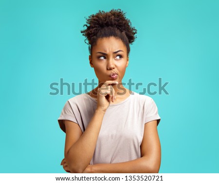 Pensive girl being deep in thoughts, raises eyebrows, curves lips, holds chin. Photo of african american girl wears casual outfit on turquoise background. Emotions and pleasant feelings concept. Royalty-Free Stock Photo #1353520721