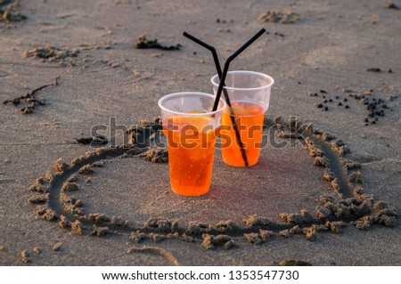 Two plastic glass with orange Aperol spritz standing inside drawn heart on sand in Zandvoort beach at sunset with black straws crossed
