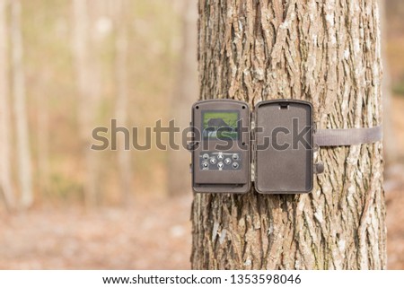 A trail camera strapped to a tree in the woods. It is open showing a photo on the screen inside. A trail camera is often used by hunters to spot deer, bear and other wildlife in the hunter's spot.