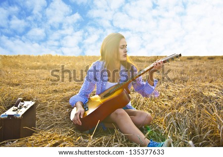 Woman chilling with accoustic guitar in wheat field on blue cloudy sky background