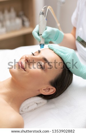 Close up of calm relaxed lady lying with her eyes closed and professional cosmetologist cleaning her skin with dermabrasion tool Royalty-Free Stock Photo #1353902882