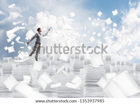 Man in casual wear keeping hand with book up while standing among flying books with cityscape on background. Mixed media.