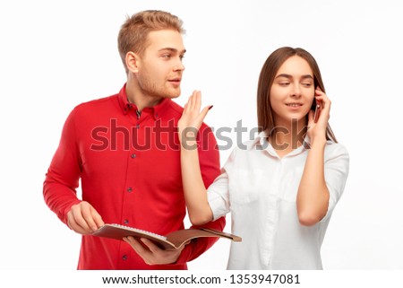 a young brunette girl in a white blouse is talking on the phone and makes a sign not to distract her young man in a red shirt with a document folder in his hands.