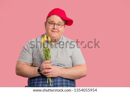 First dating when man has no amorous experience. Cute funny emotional man with plump body dressed in casual grey t-shirt and red baseball cap holding three yellow tulips over pink background.
