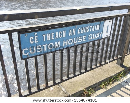 Custom House Quay road sign in English and Irish language on railings with the River Liffey in the background, in Dublin City Centre. 