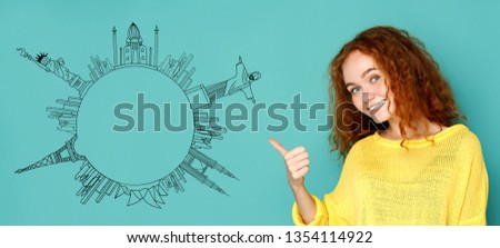 Young redhead woman with thumbs up gesture at blue background with drawn circle from world sights simbols, copy space. Travel agency concept