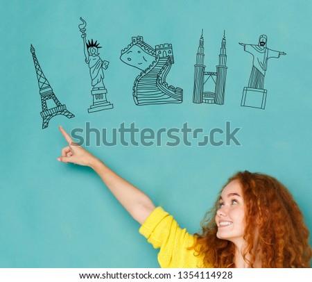 Young redhead woman pointing the finger at drawn world sights simbols on blue background. Choose your trip concept