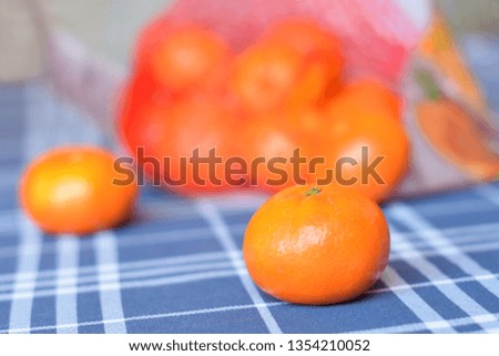 Two tangerines on a blue checkered tablecloth and a bag with more tangerines in the background