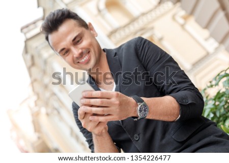 Young stylish man wearing suit standing outdoors on the city street browsing social media on smartphone close-up smiling cheerful blurry