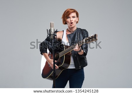 Portrait of a young woman musician with an acoustic guitar. Sing a song on studio microphone on a gray background. She wears black leather jacket and plays rock and roll loudly. Space for text
