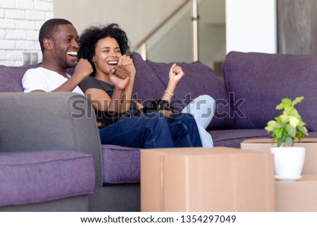 Cheerful black couple sitting on couch in living room celebrating relocation at new modern first house family feels excited and proud raising hands in yes gesture. Happy homeowners, moving day concept Royalty-Free Stock Photo #1354297049