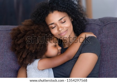 Close up biracial family portrait loving mother and little daughter sitting on couch at home hugging with closed eyes. Love, new mom for adopted child, warm relationships, caring elder sister concept Royalty-Free Stock Photo #1354297568