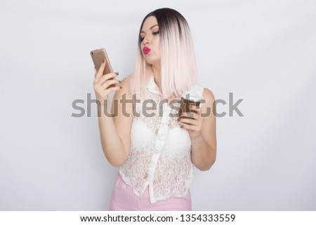 Image of cute beautiful young blonde woman in white shirt posing over white background drinking coffee and using her shartphone