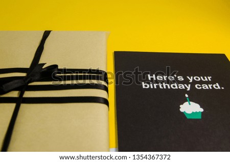 A handmade gift in brown wrapping paper and with a black ribbon.  A present and a postcard for birthday on a yellow background close up view. Texture background of a holiday preparations.