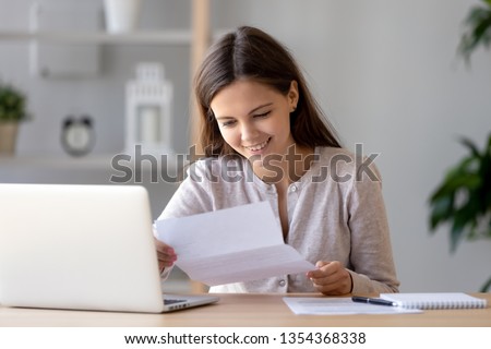 Young woman sitting at desk near laptop holding in hands paper official document about hiring recruited in company good post, bank statement financial success, college scholarship test passed concept Royalty-Free Stock Photo #1354368338