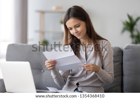 Young attractive woman sitting on sofa at home holds paper reading letter feels glad computer and notepad near on table. Student receive scholarship information or female getting loan approval concept Royalty-Free Stock Photo #1354368410