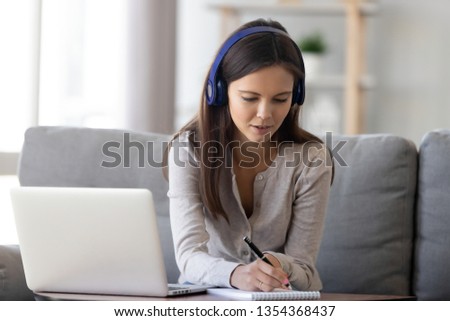 Focused young woman sitting on couch wearing headphones holding pen writing thoughts notes on notepad listening lesson lecture or webinar learning via internet use pc, improve language skills concept Royalty-Free Stock Photo #1354368437
