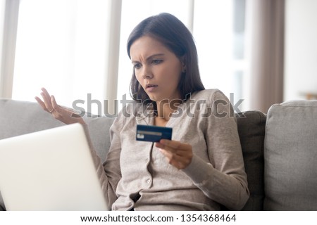 Confused woman sitting on couch holds credit card use laptop looking at device screen having debt problems, transaction failed, money withdraw impossible, insecure online payment or scam fraud concept Royalty-Free Stock Photo #1354368464