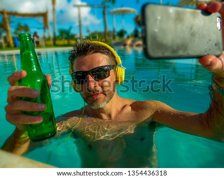 young happy and attractive man taking selfie picture with mobile phone drinking beer and listening to music at tropical resort swimming, pool enjoying Summer holidays relaxed and indulged