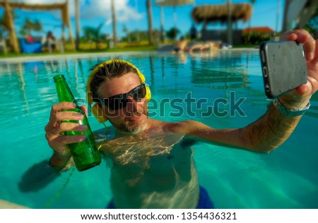 young happy and attractive man taking selfie picture with mobile phone drinking beer and listening to music at tropical resort swimming, pool enjoying Summer holidays relaxed and indulged