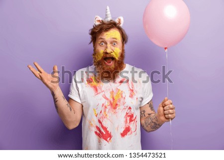 Emotional male has image of unicorn, gestures actively, has surprised expression, dirty face and t shirt, exclaims emotionally, holds balloon, entertains guests on party, suggests creative contest