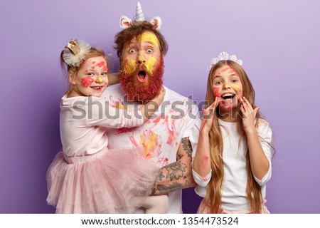 Emotional father with thick ginger beard, carries small child on hands, spends spare time with two daughters, have dirty faces with oil paints, draw picture, prepare for party, isolated on purple wall
