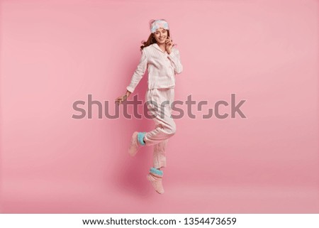 Good morning! Positive young European woman in nightwear, eyemask, jumps in air, has glad expression, has energy after awakening, isolated over pink background. People, bed time and rest concept Royalty-Free Stock Photo #1354473659